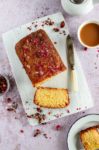 Lemon and Rose Drizzle Cake laid on a chopping board surrounded by dried rose petals. There is a knife on the chopping board and a cup of tea to the side of the board
